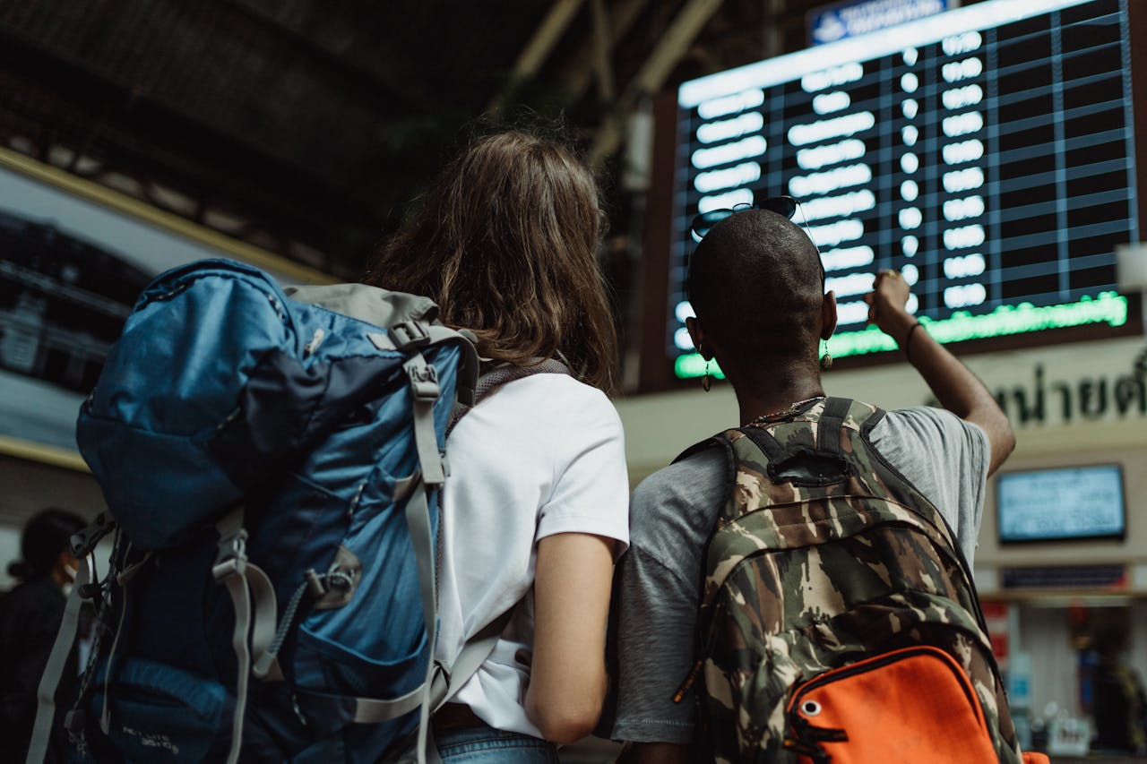 Two backpackers checking the digital board at a train station for travel information.